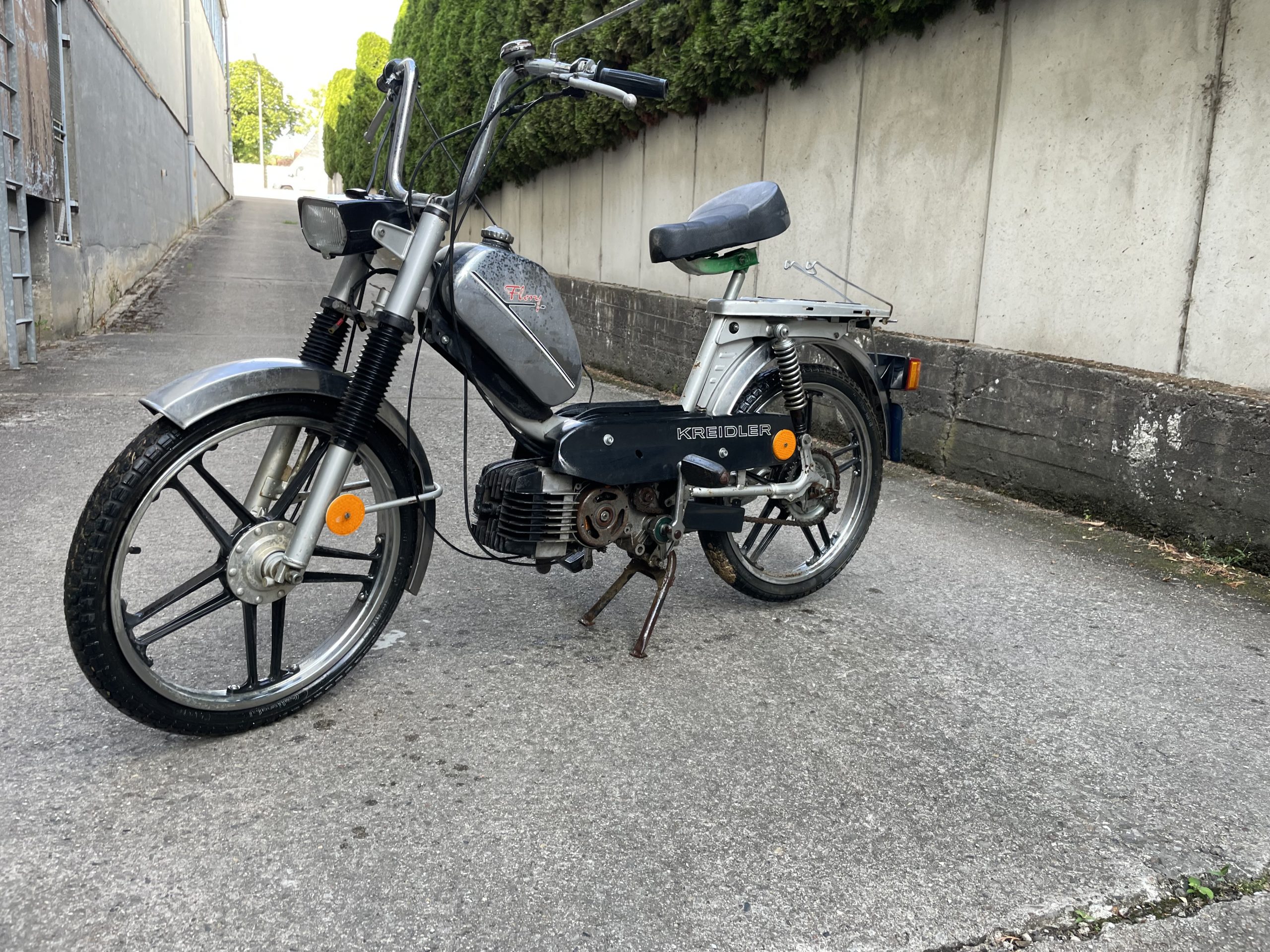 Klassisches schwarzes Moped mit Speichenrädern und Gepäckträger, geparkt in einer schmalen Gasse neben einer Betonmauer