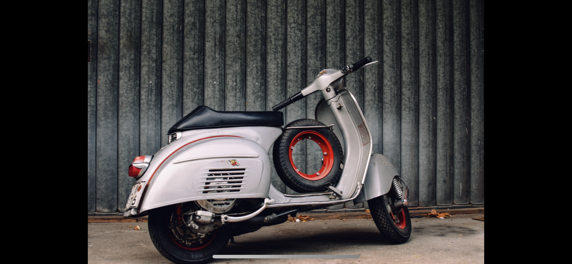 Silberne klassische Vespa mit schwarzem Sitz und roten Akzenten, geparkt vor einem grauen, gerillten Metalltor.