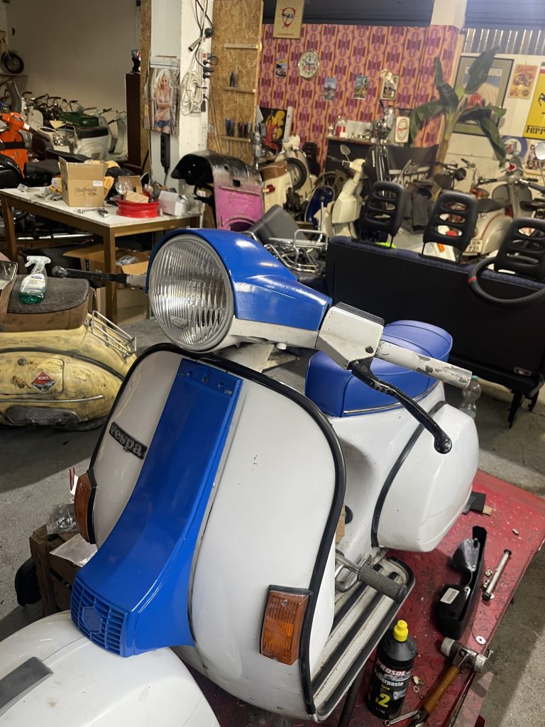 Restaurierte weiße Vespa mit blauer Verkleidung in einer Werkstatt, umgeben von weiteren Rollern, Mopeds, Ersatzteilen und Werkzeugen.
